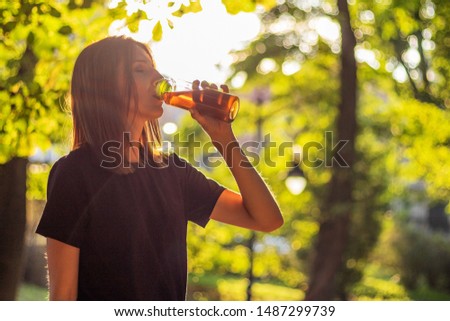 Beautiful fitness athlete woman are drinking iced tea after workout exercising on sunset evening in park. Outdoor portrait with sun glares. Royalty-Free Stock Photo #1487299739