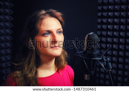 Woman in the red dress is behind the microphone. The singer looks forward to the recording Studio. Girl with blue eyes on a black background in misic studio.