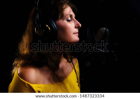 A young woman wearing headphones on a black background next to the microphone, copy space. Girl singer in yellow clothes at the recording Studio. Female portrait, close-up.