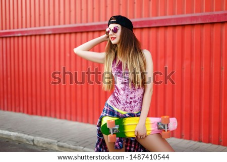 Beautiful girl in shorts, a T-shirt, glasses and a cap poses with a skateboard on the background of a red wall. Summer rest