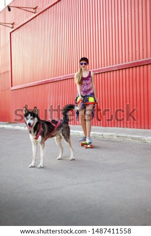 Beautiful girl in a cap and glasses skates on a skateboard with a husky dog on a background of a red wall. Summer rest