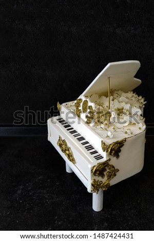 The handmade cake in the shape of a white piano in a classic style with an open lid, decorated with gold flowers and patterns. stands on a dark marble background with open space