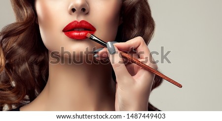 Makeup artist applies  red lipstick  . Beautiful woman face. Hand of make-up master, painting lips of young beauty  model girl . Make up in process
