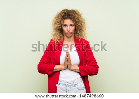 Young blonde woman with curly hair over isolated green background keeps palm together. Person asks for something
