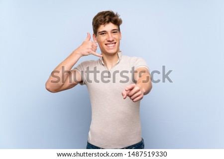 Handsome young man over isolated blue wall making phone gesture and pointing front