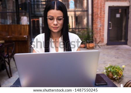 Woman skilled copywriter using applications on laptop computer, sitting in coffee shop outdoors. Female in fashionable glasses checking e-mail on notebook gadget,resting in cafe outside