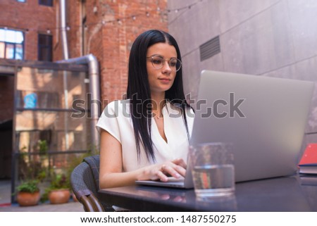 Fashionable hipster girl in spectacles connected to public wifi via pc laptop computer, sitting in coworking space outdoors. Female skilled magazine editor reading news via notebook, resting in cafe