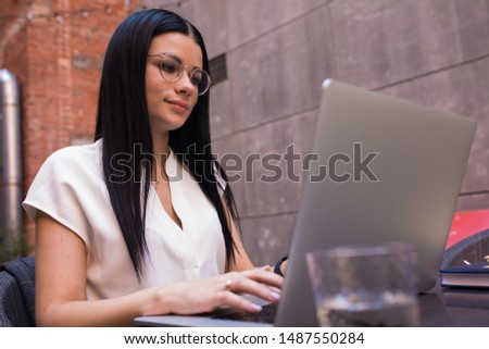 Attractive woman in stylish glasses having online training course via portable laptop computer while sitting outdoors in coworking space. Female in spectacles online booking via notebook gadget
