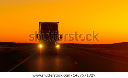 CLOSE UP LENS FLARE: Semi truck driving and hauling goods on empty highway across the Great Plains in golden morning. Freight delivery truck transporting cargo on interstate freeway at stunning sunset Royalty-Free Stock Photo #1487571929
