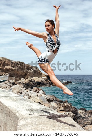 The gymnast girl splits in the air. Beautiful young gymnast woman performing an element of rhythmic gymnastics, jumping, making a jump in the air, dancing near the sea. Greece.