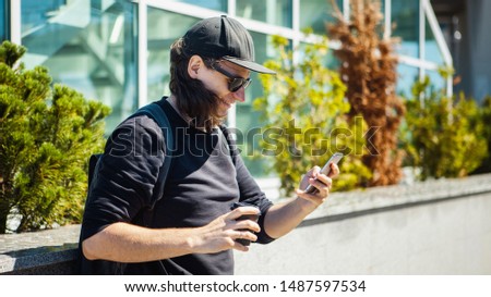 A man in black glasses, a black baseball cap and shoots coffee on the street on a smartphone. Side view. Paparazzi. Private investigations.