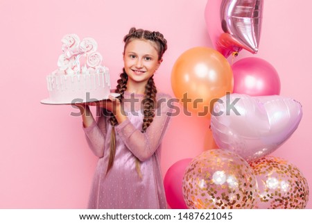 A teenage girl with balloons holds a cake in her hands, on a pink background. Birthday concept