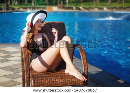 Young woman in a black bathing suit and beach hat sits in a deckchair of artificial rattan near the pool with blue water in the resort hotel.