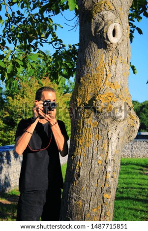 Man in black T-shirt taking pictures with a camera, hidden behind a tree trunk. In nature with vegetation and countryside in the background.