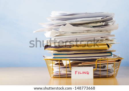 An old yellow wireframe filing tray, piled high with documents and folders, on a light wood veneer desk against light blue background. Royalty-Free Stock Photo #148773668