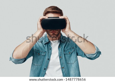 Handsome young man using virtual reality simulator while standing against grey background