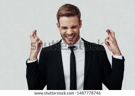 Handsome young man in formalwear keeping fingers crossed and eyes closed while standing against grey background