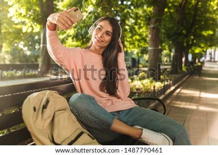Smiling young girl listening to music with headphones while sitting on a bench at the city park and taking a selfie mobile phone