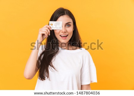 Image of beautiful brunette woman wearing basic clothes smiling and holding credit card isolated over yellow background