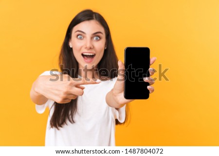 Image of gorgeous brunette woman wearing basic clothes rejoicing and pointing finger at cellphone isolated over yellow background