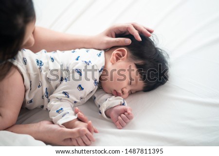 Top view of Asian mother is touching her cute sleeping newborn baby head on bed. She smiling and putting her hand a warm touching on kid hair with love in white bedroom at home. Happy family concept.