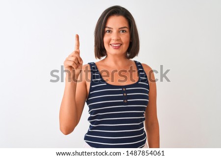 Young beautiful woman wearing blue striped t-shirt standing over isolated white background showing and pointing up with finger number one while smiling confident and happy.
