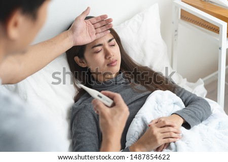 Sick day at home in winter season. Young asian woman lying on the bed have a fever and her husband checking thermometer and measure body temperature. Royalty-Free Stock Photo #1487854622