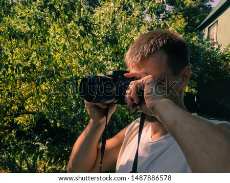 A man with a camera photographs against the backdrop of green vegetation on a summer sunny day.