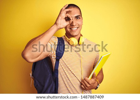Young student man wearing headphones and backpack over yellow isolated background with happy face smiling doing ok sign with hand on eye looking through fingers