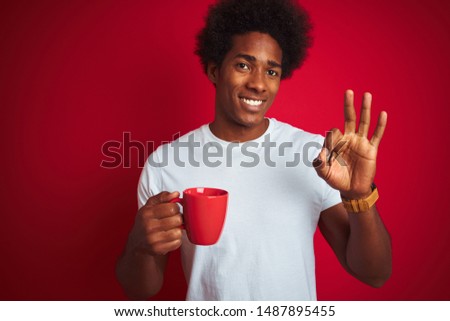 Young african american man drinking a cup of coffee standing over isolated red background doing ok sign with fingers, excellent symbol