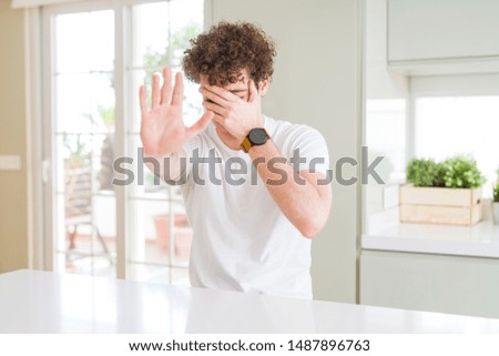 Young handsome man wearing white t-shirt at home covering eyes with hands and doing stop gesture with sad and fear expression. Embarrassed and negative concept.