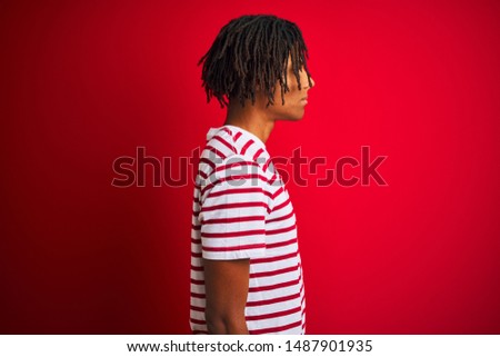 Young afro man with dreadlocks wearing striped t-shirt standing over isolated red background looking to side, relax profile pose with natural face and confident smile.