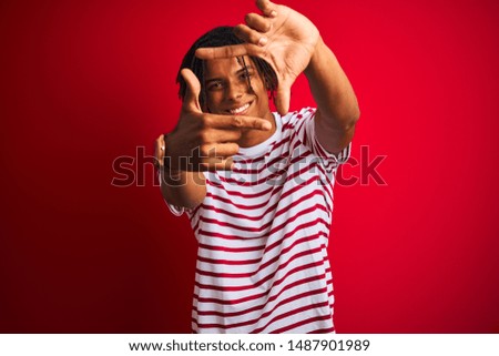 Young afro man with dreadlocks wearing striped t-shirt standing over isolated red background smiling making frame with hands and fingers with happy face. Creativity and photography concept.