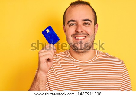 Young man holding blue credit card standing over isolated yellow background with a happy face standing and smiling with a confident smile showing teeth