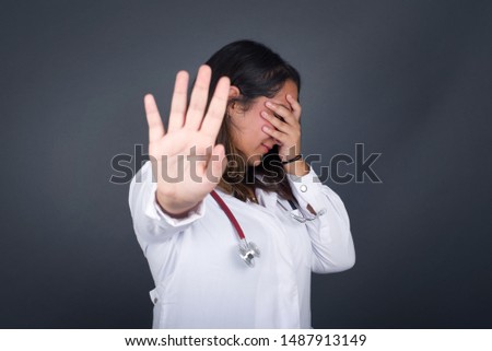 People, body language. Young European doctor woman covers eyes with palm and doing stop gesture, tries to hide from everybody. Don't look at me, I don't want to see, feels ashamed or scared.