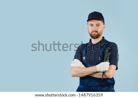 Confident service worker with hands crossed holding adjustable spanner in the hand. He is dressed in dark blue overalls, shirt, cap and cotton work gloves. Royalty-Free Stock Photo #1487916359