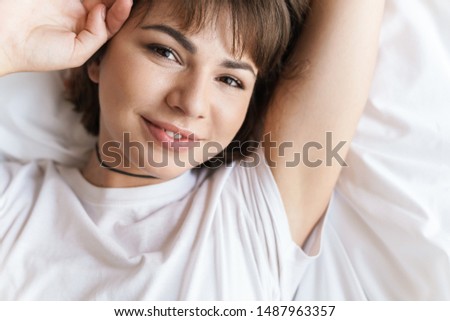 Photo of a cheerful young beautiful woman lies indoors at home on bed looking camera.