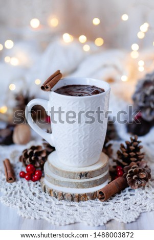 Composition with white mug with hot chocolate with spices. Decor rustic style with natural elements: cones, red berries, wood, cinnamon sticks, nuts. Christmas lights on, knitted plaid, cozy morning