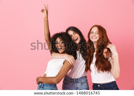 Three cute excited young girlfriends wearing casual outfits standing isolated over pink background, hugging