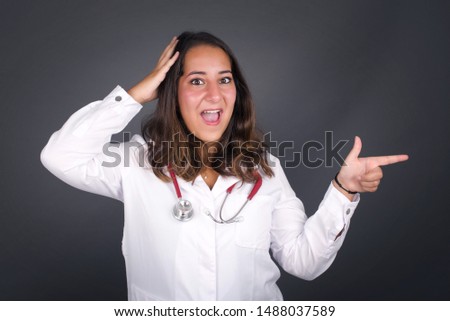 Portrait of young doctor woman with shocked facial expression, showing something amazing on blank space, one hand on her head and pointing with forefinger. OMG concept.