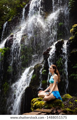 Yoga lifestyle. Young Caucasian woman sitting on the rock, meditating, practicing yoga, pranayama with gyan mudra at waterfall. Yoga retreat. Banyu Wana Amertha waterfall Wanagiri, Bali, Indonesia