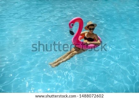 Woman in swimsuit and hat swimming with inflatable flamingo ring in the water pool, enjoying vacations during the summer time