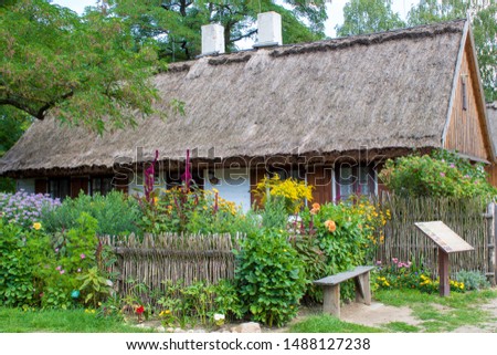 Authentic reconstructed house of the last century with a thatched roof, patio, wicker fence, flower beds on a summer day. Architecture of the early 20th century in the countryside of Europe.