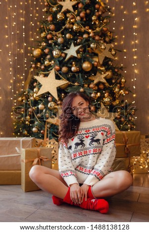 A beautiful woman in a white sweater is sitting waiting for a holiday near the New Year tree decorated with golden garlands and Christmas decorations. The idea and concept of Christmas and New Year