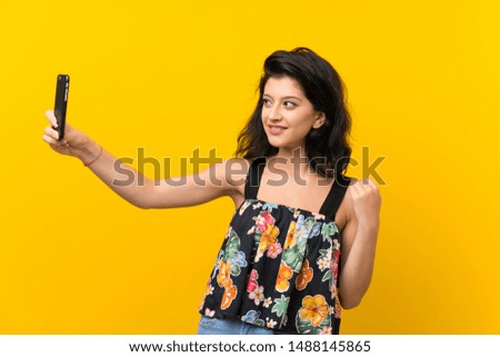 Young woman over isolated yellow background making selfie with cellphone