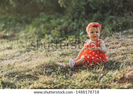 Little girl at red dress sitting on the grass at field. Summer, sunset time