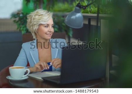 a woman with blond hair in a business suit sits at a table working on a laptop and writes in a diary, drinks coffee