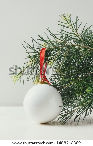 White porcelain Christmas ball with red ribbon on fir tree on white marble table. Modern Christmas concept.