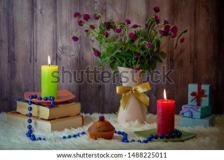 Red and green burning candles, books, a violet bouquet of wildflowers in a vase, gift boxes decorated with bows, long blue beads, a delicious cupcake on a white fluffy bedspread, wooden background.