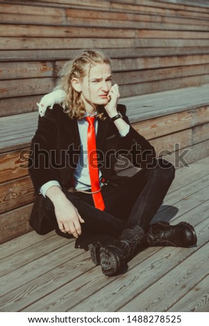 Attractive young man with long blonde hair in a black suit and red tie, rats running around his body. His face expresses displeasure.